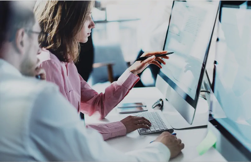 Two colleagues work together at a computer, with one pointing at the screen.