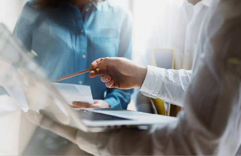 Two people collaborate at a laptop, with one pointing at the screen using a pencil.