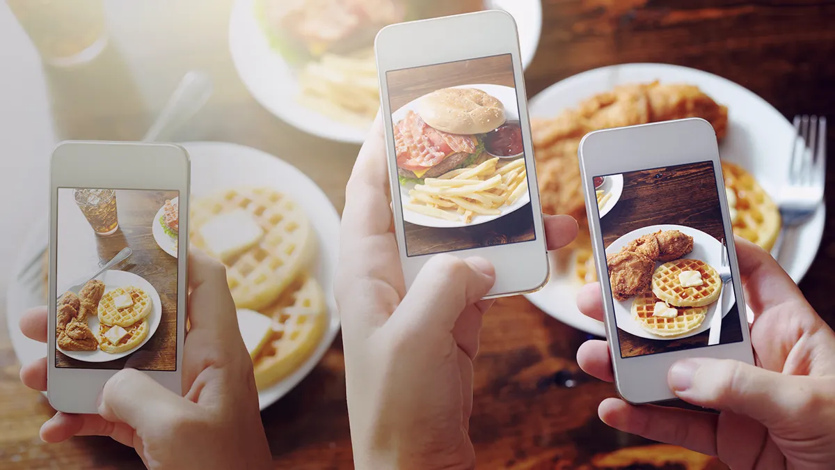The photo depicts three hands, with a smartphone in each hand; the smartphone screen shows images of food - two of the hands show pancakes, the third shows a hamburger with fries; below the hands, in the background, you can see a slightly blurry table top and the same food shown on the phones' screen. The photo is the cover graphic for a success story by Transition Technologies PSC titled Augmented Reality technology applied in gastronomy.