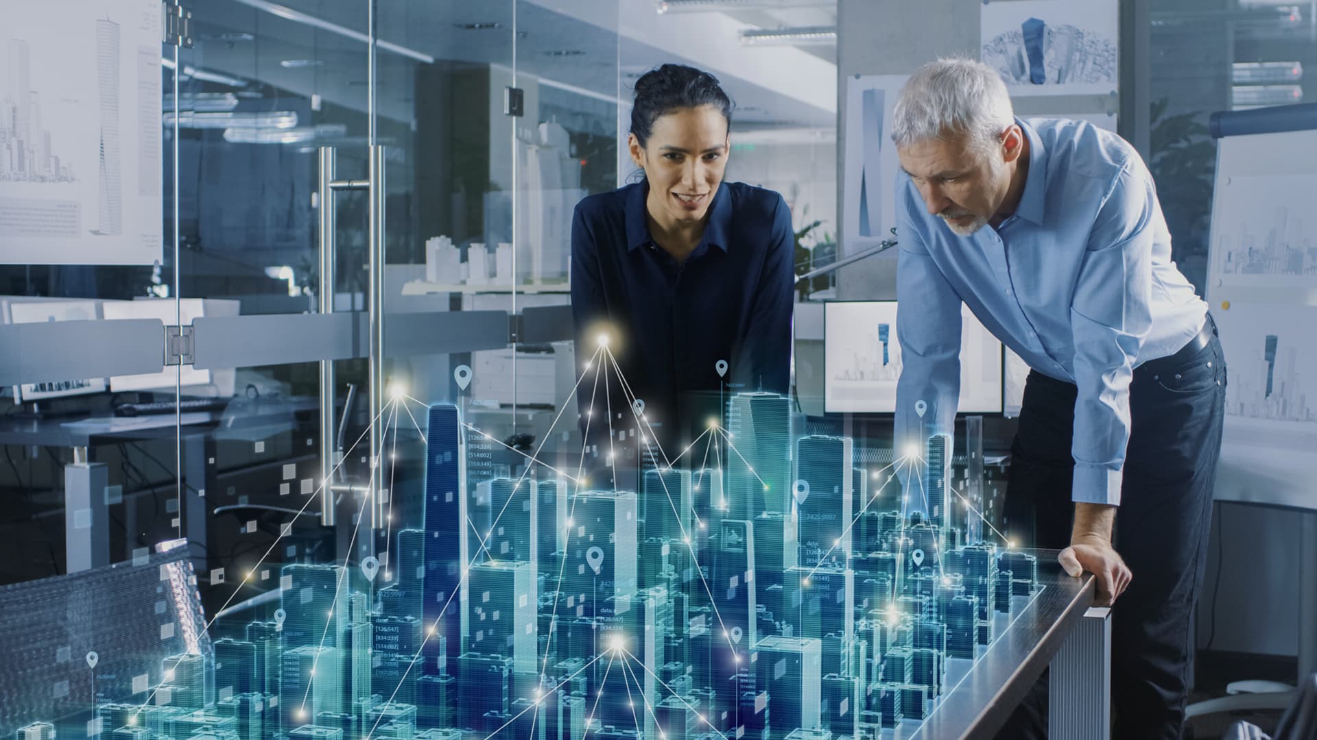 The photo shows an office room, glass doors and walls, boards with charts in the background. There is a man and a woman in the room, both of them are slightly bent over a long table, on which one can see the city displayed in a three-dimensional format, along with lines and highlighted points in some places. From the context, it appears that they are designers using modern software using Augmented Reality technology. The photo is the cover graphic for a Success Story from Transition Technologies PSC and appears as an illustration of an entry titled: AR Holographic Gallery of Lodz city.