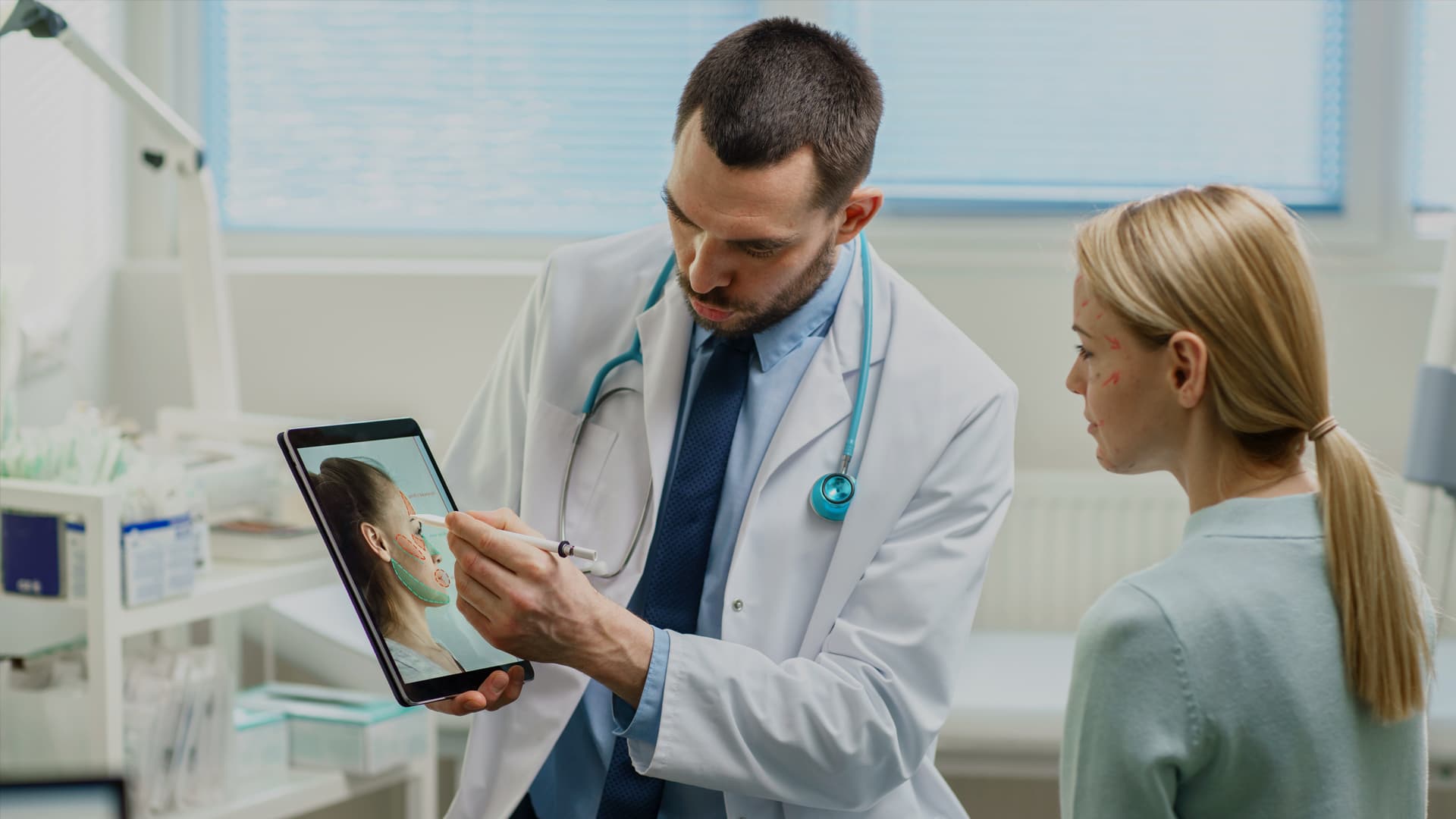 The photo shows a doctor's office and his patient; the doctor is dressed in a white smock with a stethoscope slung across his neck, and in his hands he holds a tablet that shows the patient's face with several areas marked. The man points to the patient's forehead in the photo displayed on the tablet. The patient is facing the screen. From the context, it appears to be a modern aesthetic medicine office. The photo is the cover graphic for Transition Technologies PSC's Success Story and appears as an illustration of an entry titled: The application of Augmented Reality in esthetic medicine and plastic surgery.