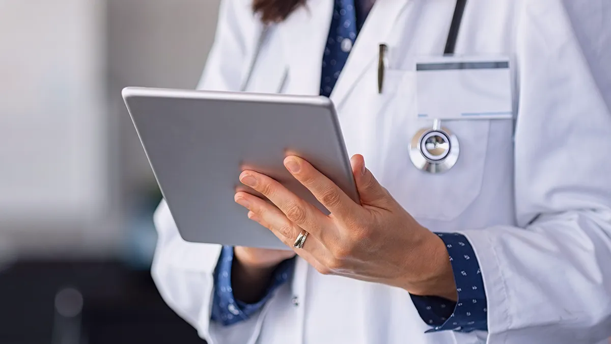 Das Foto zeigt den Torso einer Frau in einem Arztkittel, mit einem Stethoskop um den Hals und einem Abzeichen auf der linken Brust. Im Vordergrund ist die linke Hand der Frau zu sehen, die ein Tablet hochhält. Aus dem Kontext heraus scheint es sich um eine Ärztin zu handeln, die einige Daten auf dem Bildschirm eines mobilen Geräts überprüft. Das Foto ist die Titelgrafik für eine Erfolgsgeschichte von Transition Technologies PSC mit dem Titel: Digitalisierung von gesundheitswesen – IT im regierungsprojekt.