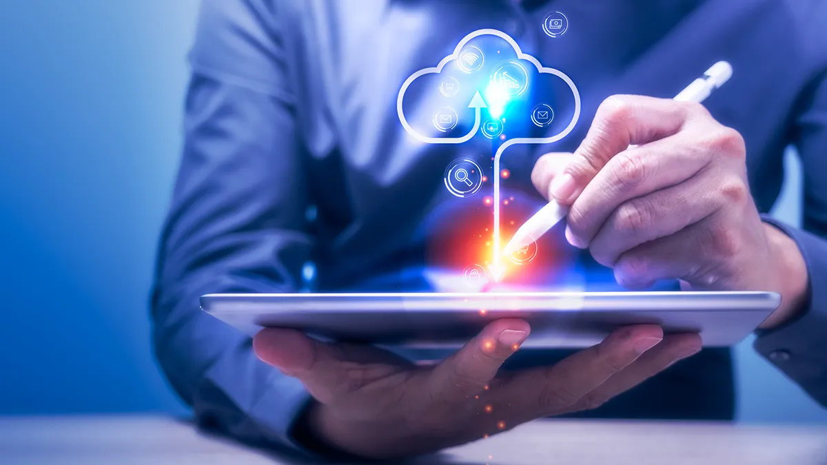 Eine Person im Business-Anzug hält ein Tablet mit Stift in der Hand. Auf dem Tablet-Bildschirm ist eine Wolkengrafik zu sehen, die von verschiedenen Symbolen umgeben ist, darunter Währungssymbole und nach oben zeigende Pfeile, die Wachstum suggerieren. Der Hintergrund ist mit einem blauen Farbton verschwommen, wodurch die Aufmerksamkeit auf die Hände, das Tablet und die grafische Darstellung der Cloud-Dienste gelenkt wird. Bild für einen Blog-Artikel: Wie lässt sich mit FinOps eine AWS-Cloud-Kostenoptimierung erreichen?