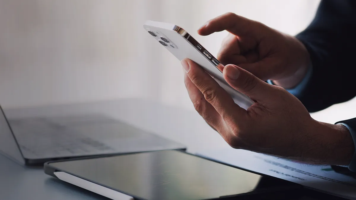 Das Foto zeigt die Hände eines Mannes, der ein weißes Mobiltelefon hält, der Rest der Figur ist außerhalb des Bildausschnitts. Der Mann ist mit einem Hemd und einer Jacke bekleidet, auf der Kommode, über die er das Telefon hält, sind ein offener Laptop und ein Tablet zu sehen; aus dem Kontext heraus sieht es so aus, als ob der Mann bei der Arbeit ist. Das Farbschema ist hell. Das Foto ist die Titelgrafik für eine Erfolgsgeschichte von Transition Technologies PSC mit dem Titel: QA in OneSpan Mobile – globale Testautomatisierung.