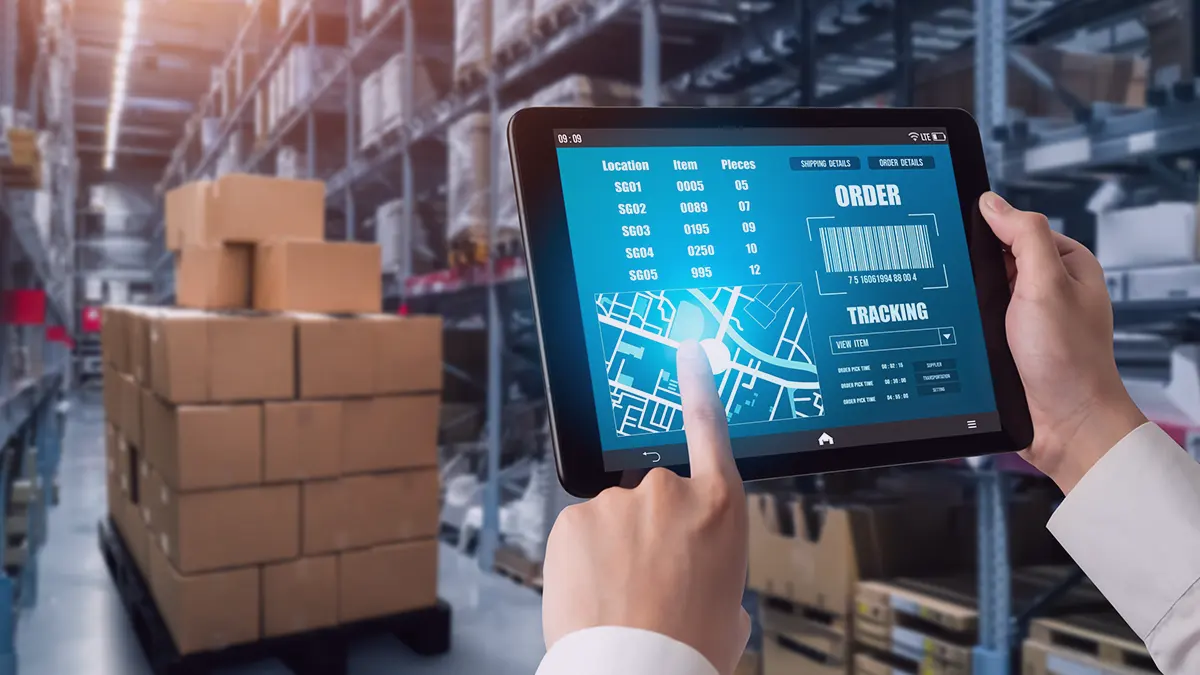 Zdjęcie stanowiące okładkę wpisu na blogu firmy Transition Technologies PSC, przedstawia tablet trzymany w dłoniach widoczny w prawym dolnym rogu, reszta postaci nie jest widoczna; na ekranie urządzenia wyświetlane są dane dotyczące zamówienia, liczy, mapa i kod QR, na drugim planie widoczna jest paleta kartonów, z kontekstu wynika, że są to towary zapakowane do wysyłki; w tle widać metalowe półki magazynowe, pełne różnych produktów.