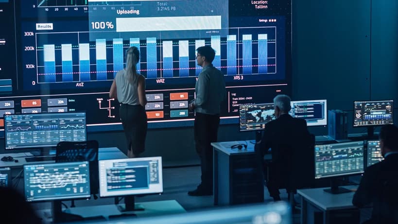 A high-tech operations center with professionals monitoring multiple data screens displaying industrial analytics, system performance metrics, and real-time data visualization. A large central display shows detailed graphs and progress indicators, highlighting operational insights and efficiency tracking.