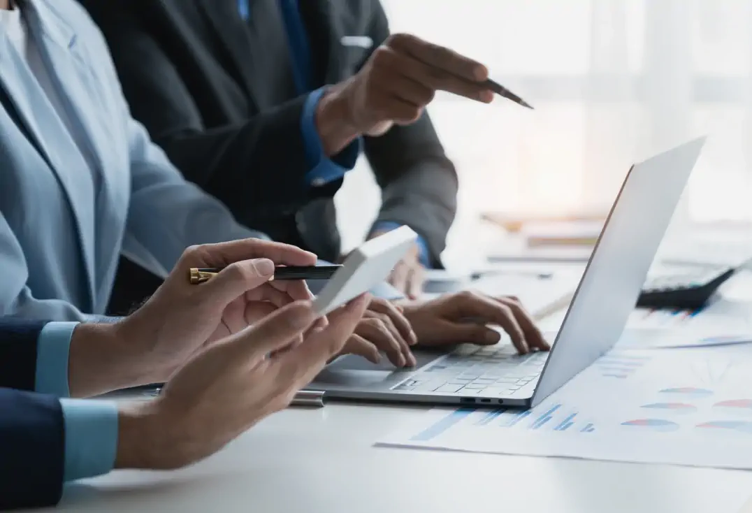 Business professionals working together at a desk with a laptop, documents, and digital devices, discussing data and strategies.