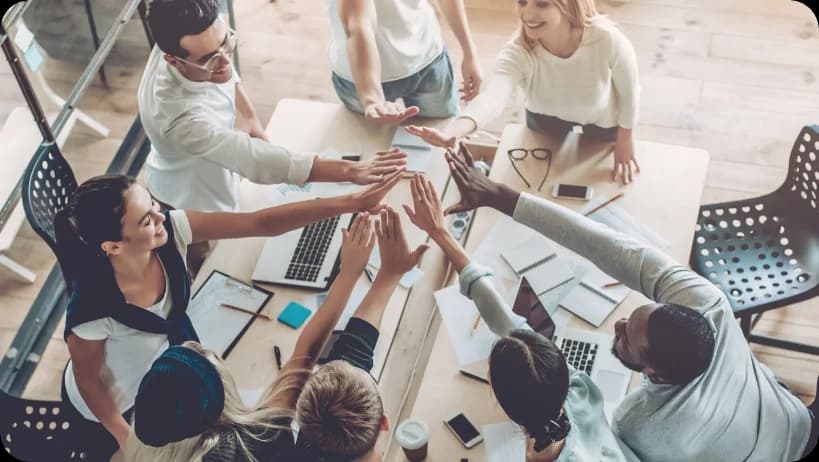 Team of colleagues in an office doing a group high five around a table with laptops and documents.