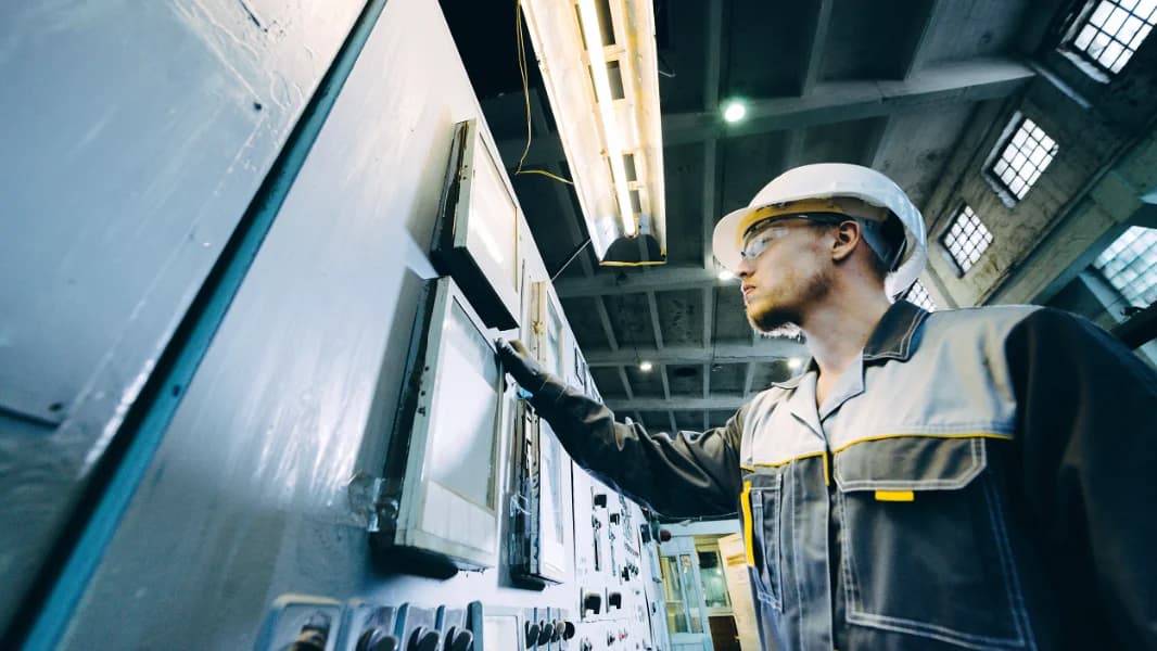 An employee wearing a helmet and a reflective vest supervises a large machine component in the production hall.