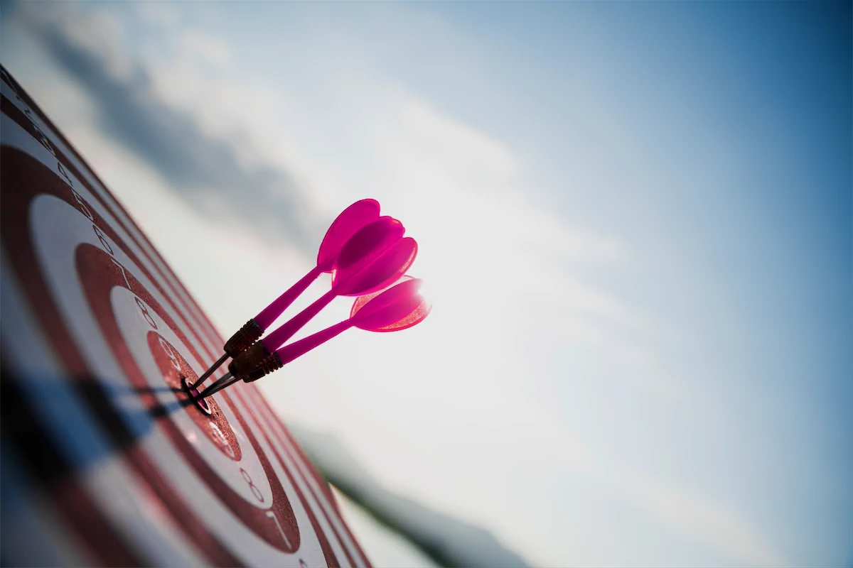 Three pink darts hitting the bullseye of a dartboard against the sky.
