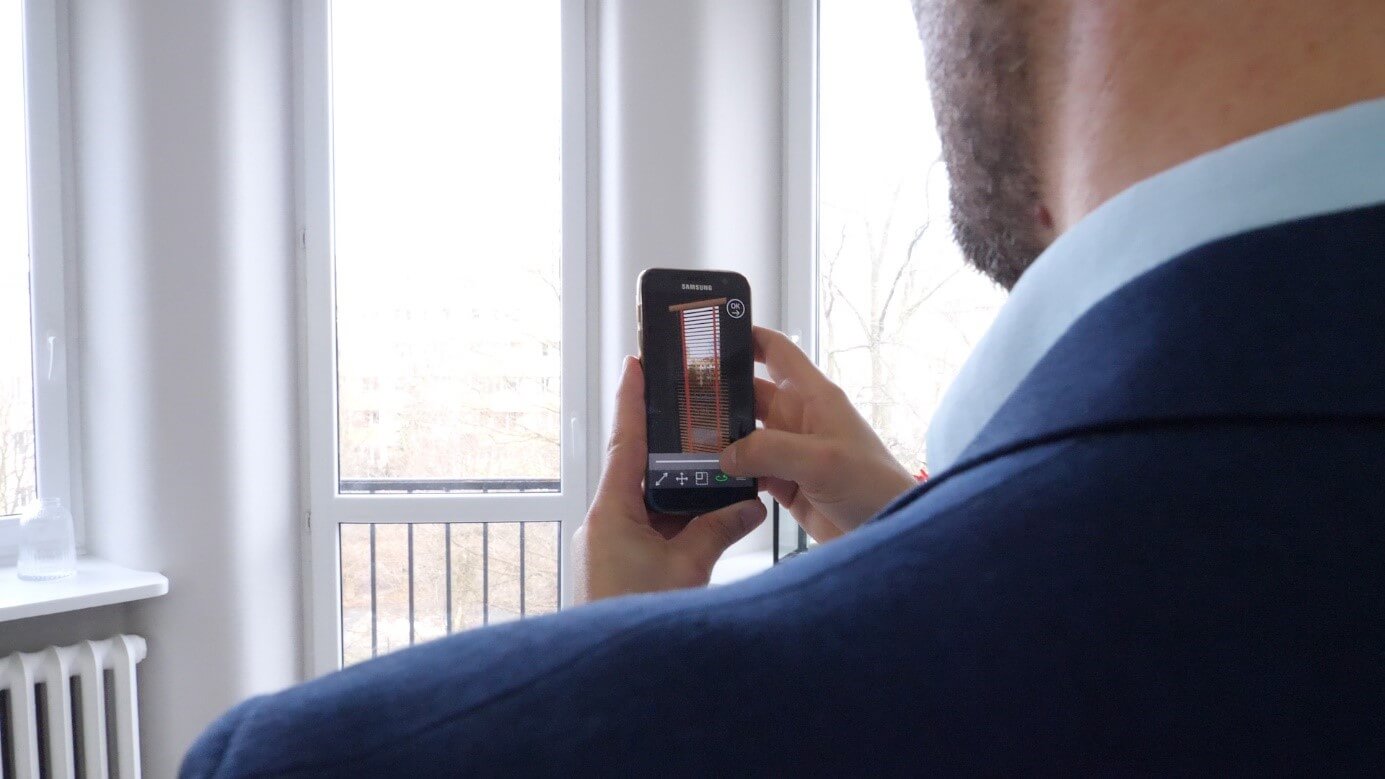 A man in a suit stands in front of a balcony window with his smartphone. His back is turned and he measures the window using an AR app. The app matches the product dimension to the window and takes measurements. 
